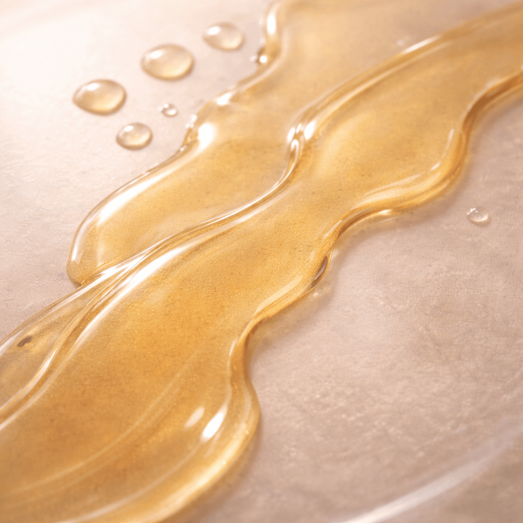 Close-up of a flowing gold liquid Silk Body Oil on a beige background